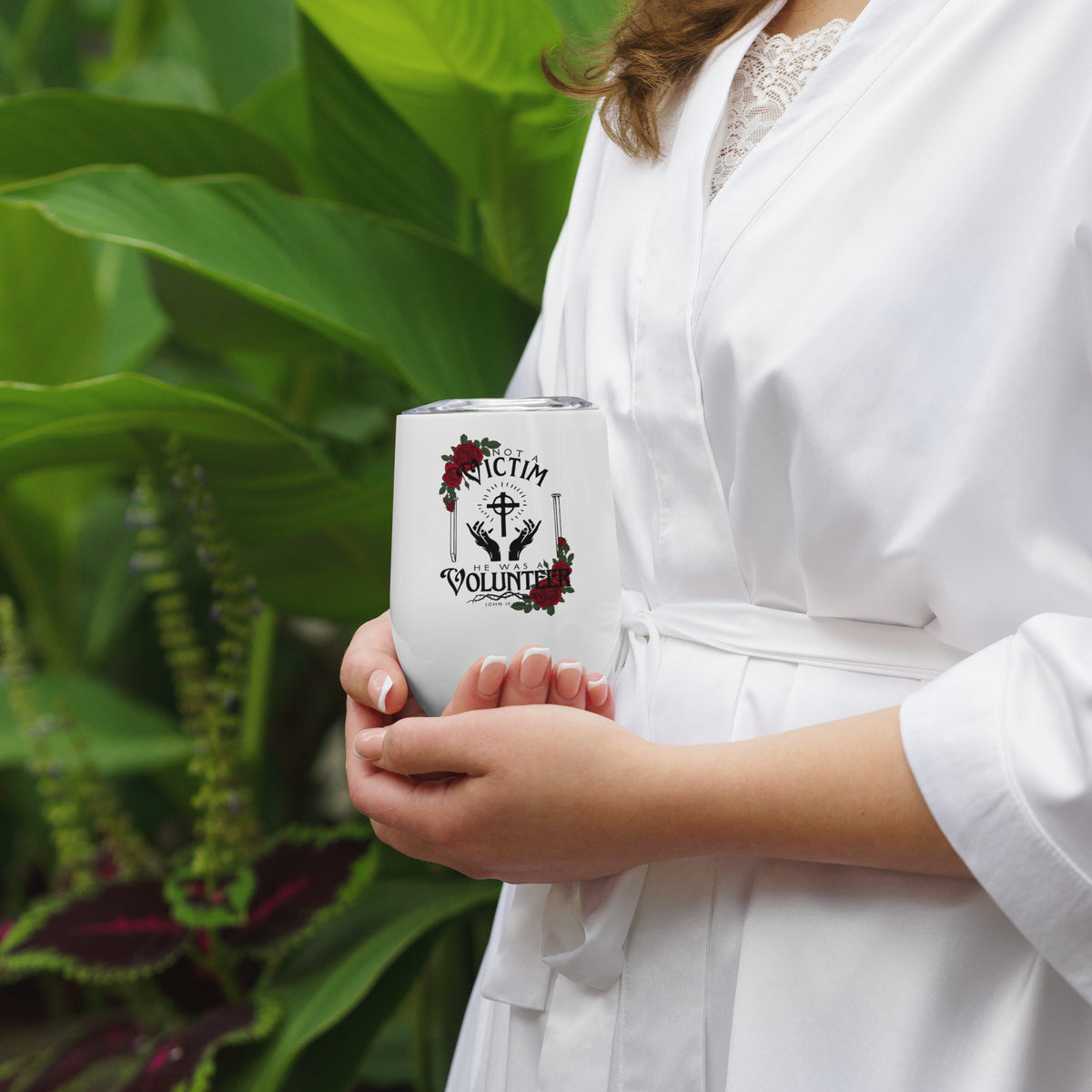 Person holding a white Not a Victim Christian wine tumbler outdoors near green plants